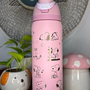 Puede incluir: Botella de agua aislada rosa con ilustraciones de Snoopy y detalles dorados. La botella tiene una tapa rosa con el texto "Lift to Unlock". Un art&iacute;culo decorativo y funcional para el uso diario.