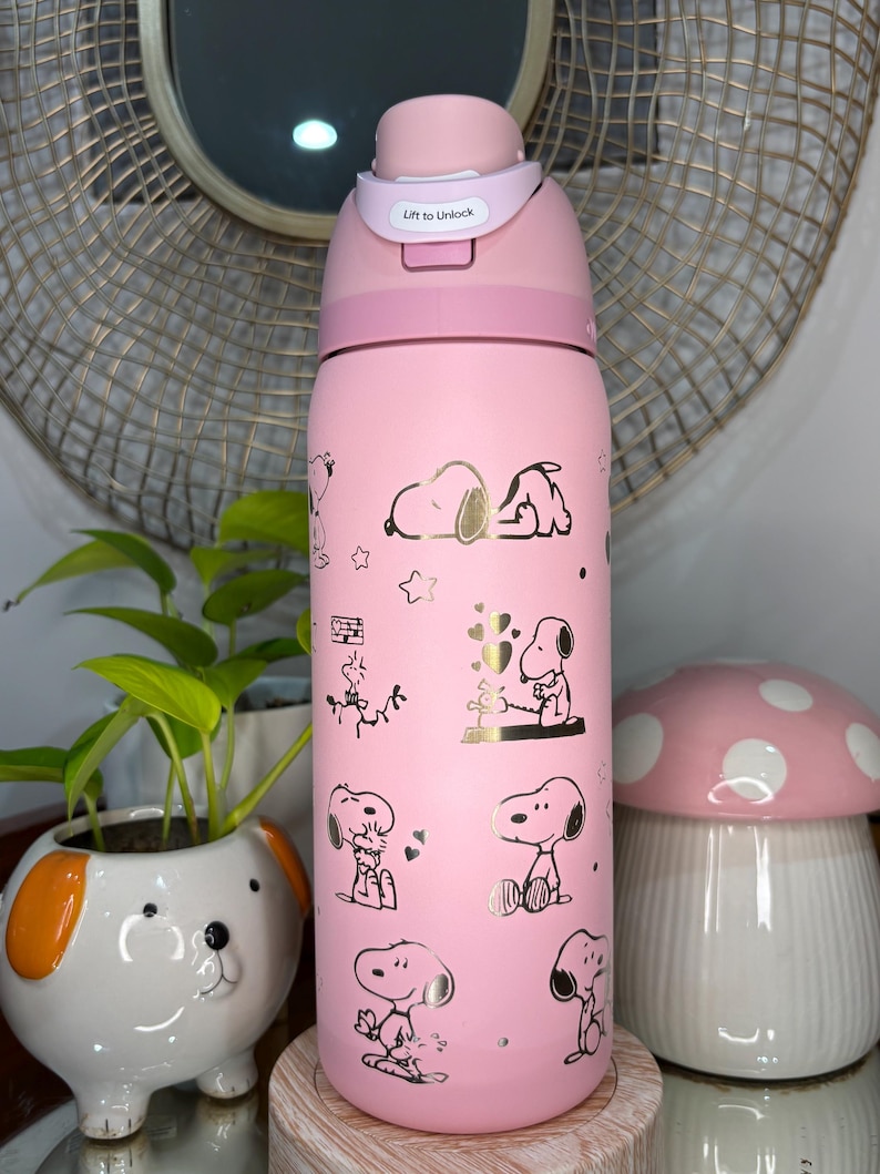 Puede incluir: Botella de agua aislada rosa con ilustraciones de Snoopy y detalles dorados. La botella tiene una tapa rosa con el texto "Lift to Unlock". Un art&iacute;culo decorativo y funcional para el uso diario.