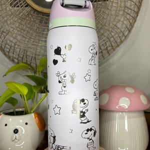 Puede incluir: Una botella de agua aislada blanca con personajes de dibujos animados de Snoopy y estrellas doradas. La botella tiene una tapa rosa, verde y blanca con asa. La botella mide aproximadamente 23 cm de alto.
