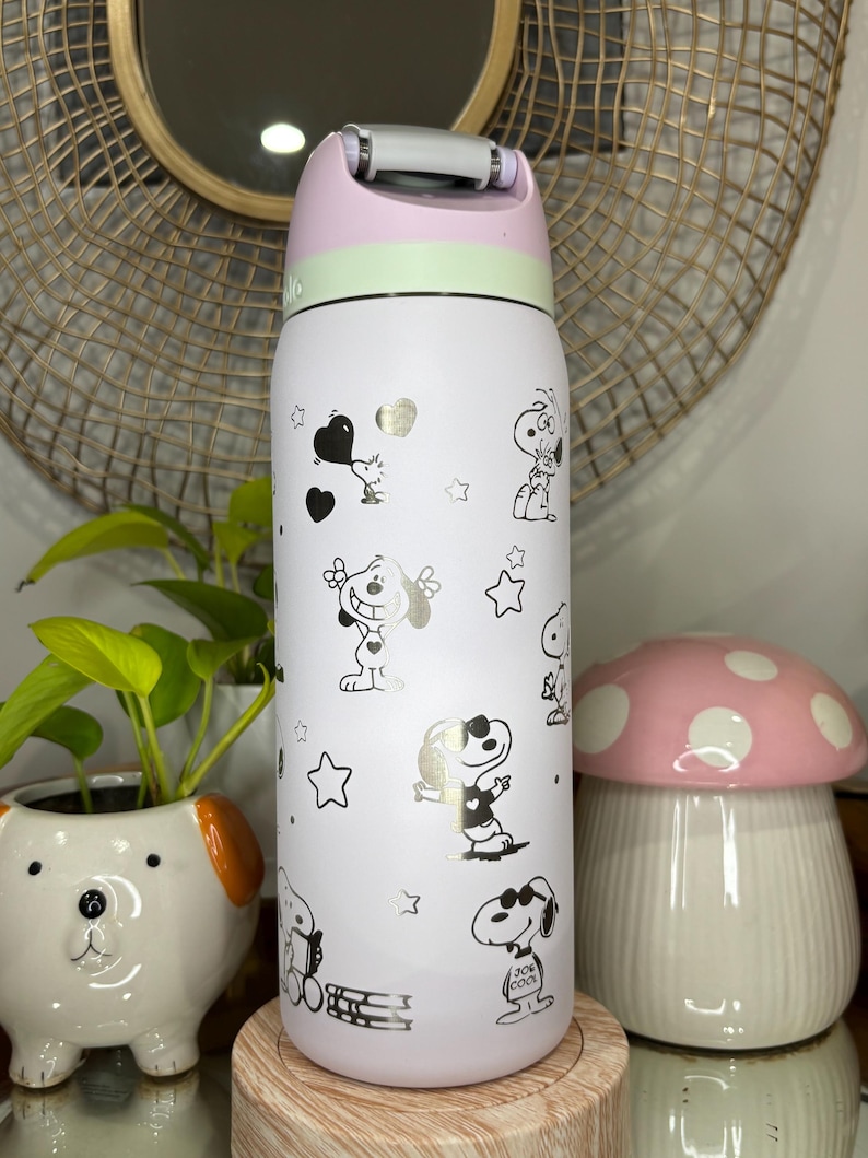 Puede incluir: Una botella de agua aislada blanca con personajes de dibujos animados de Snoopy y estrellas doradas. La botella tiene una tapa rosa, verde y blanca con asa. La botella mide aproximadamente 23 cm de alto.