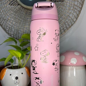 Puede incluir: Una botella de agua rosa Owala con ilustraciones doradas del personaje Snoopy. La botella tiene una tapa abatible y un asa. Un macetero blanco y naranja con forma de perro y un recipiente con forma de hongo rosa est&aacute;n en el fondo.