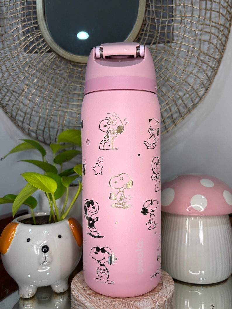 Puede incluir: Una botella de agua rosa Owala con ilustraciones doradas del personaje Snoopy. La botella tiene una tapa abatible y un asa. Un macetero blanco y naranja con forma de perro y un recipiente con forma de hongo rosa est&aacute;n en el fondo.