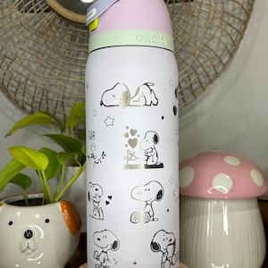 Puede incluir: Una botella de agua Owala blanca con ilustraciones de Snoopy en dorado y negro. La botella tiene una tapa rosa y verde y un asa gris. Un peque&ntilde;o macetero con forma de perro y un adorno con forma de hongo est&aacute;n en el fondo.