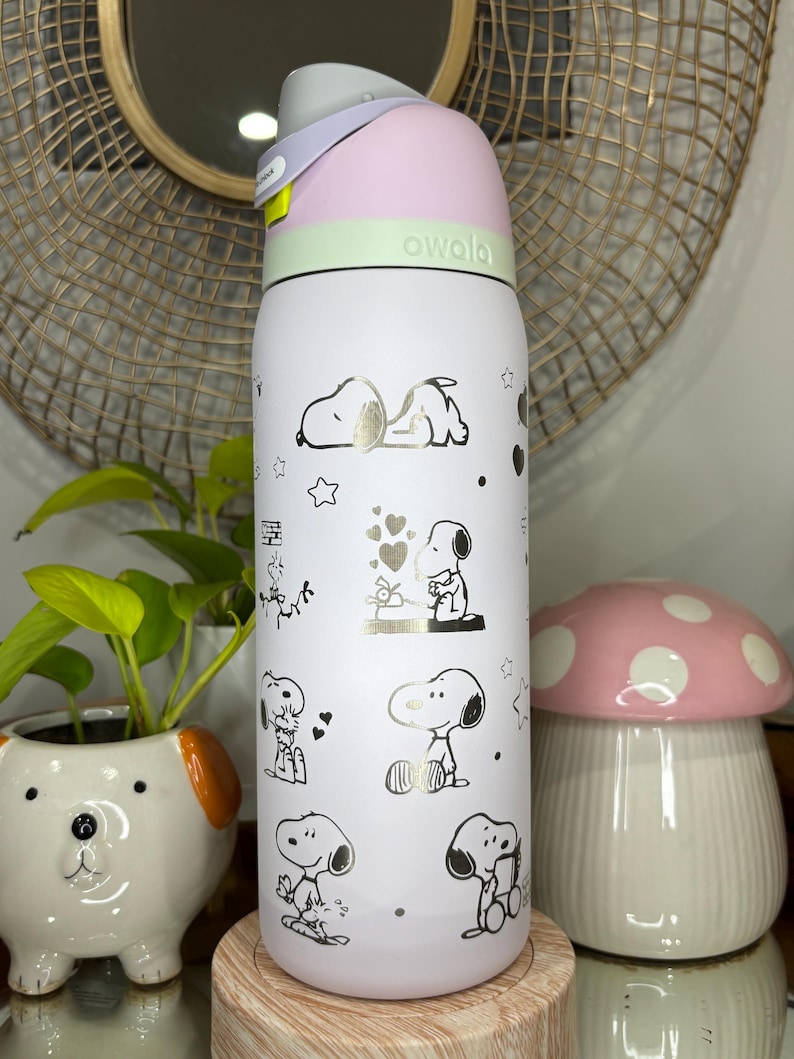 Puede incluir: Una botella de agua Owala blanca con ilustraciones de Snoopy en dorado y negro. La botella tiene una tapa rosa y verde y un asa gris. Un peque&ntilde;o macetero con forma de perro y un adorno con forma de hongo est&aacute;n en el fondo.