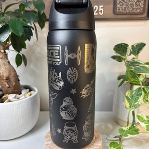 May include: Black water bottle with a flip-top lid, featuring etched Star Wars characters and phrases. The design includes the text "FORCE WITH YOU", along with images of the X-wing, BB-8, and Stormtroopers. The bottle is displayed on a wooden coaster.