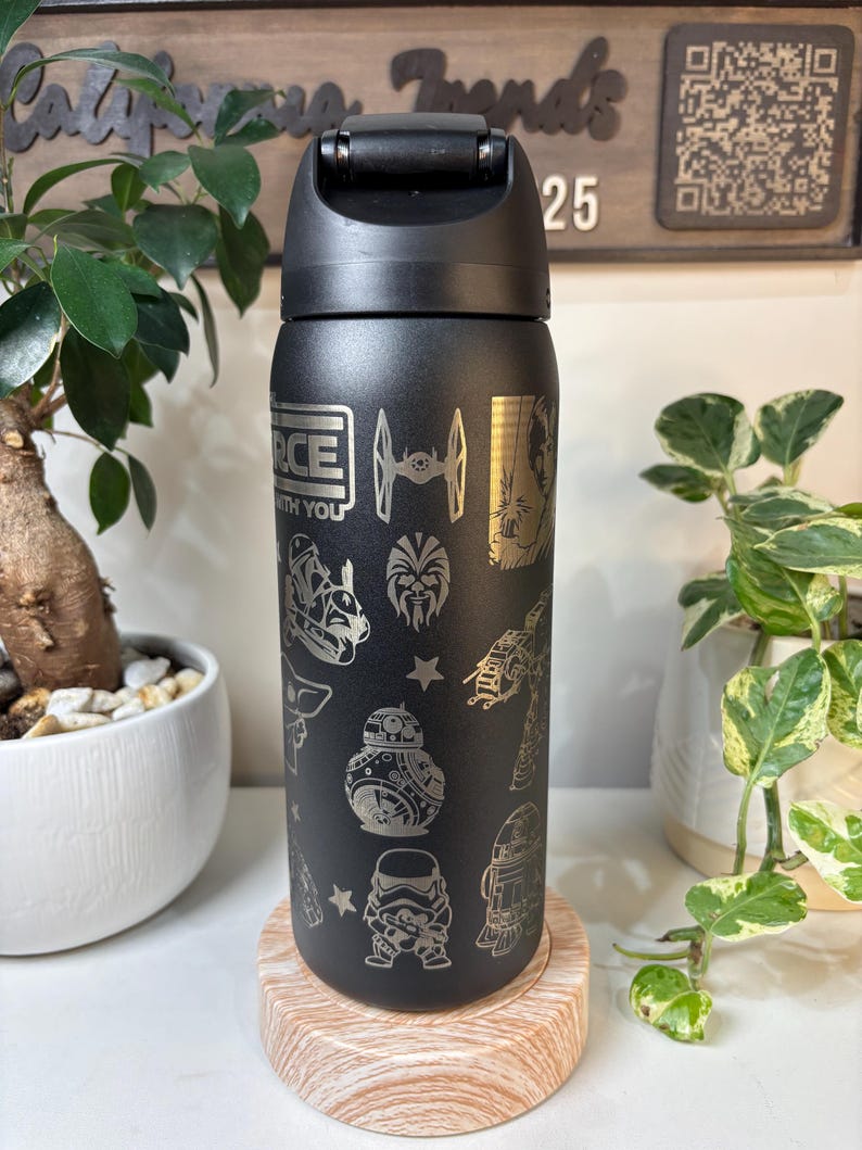 May include: Black water bottle with a flip-top lid, featuring etched Star Wars characters and phrases. The design includes the text "FORCE WITH YOU", along with images of the X-wing, BB-8, and Stormtroopers. The bottle is displayed on a wooden coaster.