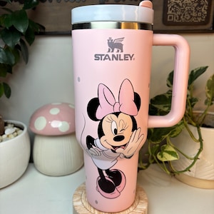 Puede incluir: Un termo de viaje Stanley rosa con un dise&ntilde;o de Minnie Mouse. El termo tiene un asa y una tapa blanca. El logotipo de Stanley es visible. El termo est&aacute; sobre un posavasos de madera. El fondo incluye plantas y un letrero decorativo.
