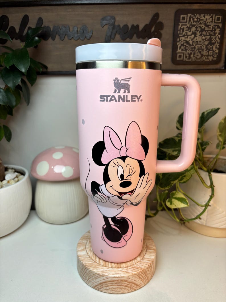 Puede incluir: Un termo de viaje Stanley rosa con un dise&ntilde;o de Minnie Mouse. El termo tiene un asa y una tapa blanca. El logotipo de Stanley es visible. El termo est&aacute; sobre un posavasos de madera. El fondo incluye plantas y un letrero decorativo.
