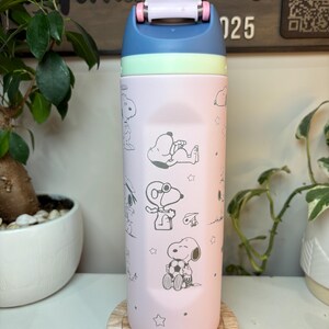May include: A pink and blue insulated water bottle featuring Snoopy and other Peanuts characters. The bottle has a blue lid with a white handle and a light green band. The design includes stars and various Snoopy illustrations.