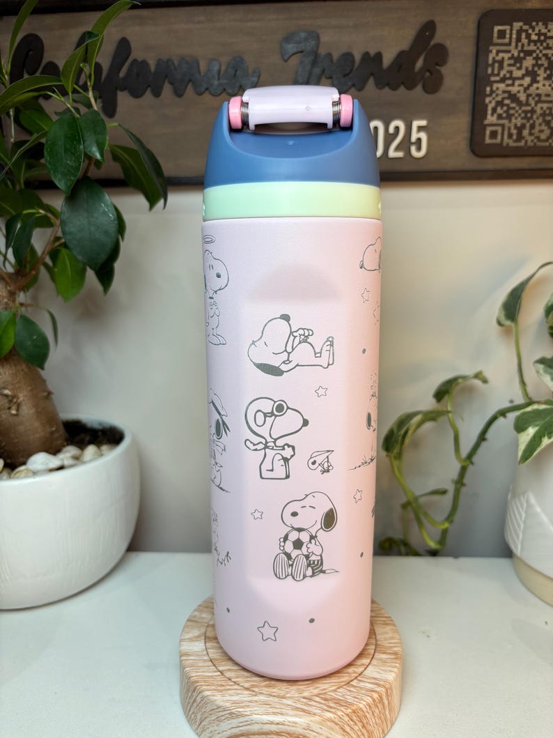 May include: A pink and blue insulated water bottle featuring Snoopy and other Peanuts characters. The bottle has a blue lid with a white handle and a light green band. The design includes stars and various Snoopy illustrations.