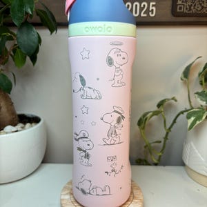 May include: A pink Owala water bottle featuring Snoopy illustrations in various poses. The bottle has a blue and white lid with a pink handle. The word "owala" is printed in green on the bottle. The bottle is sitting on a wooden coaster.