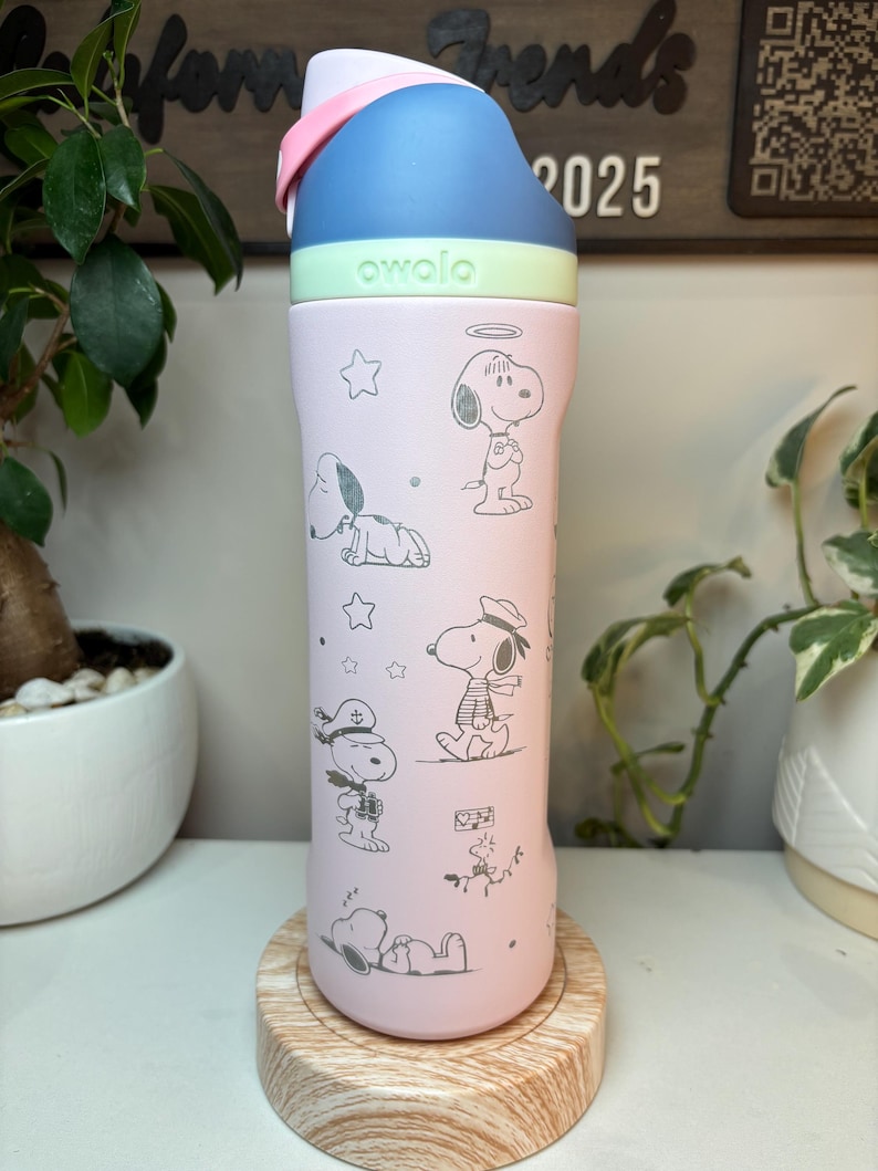 May include: A pink Owala water bottle featuring Snoopy illustrations in various poses. The bottle has a blue and white lid with a pink handle. The word "owala" is printed in green on the bottle. The bottle is sitting on a wooden coaster.