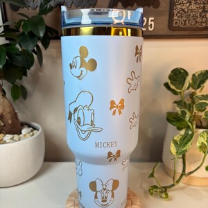 Puede incluir: Un vaso azul claro con dise&ntilde;os dorados de Mickey Mouse, Donald Duck y Minnie Mouse, junto con lazos y guantes. El vaso tiene un borde dorado y una tapa azul transparente. La palabra "Mickey" est&aacute; impresa.