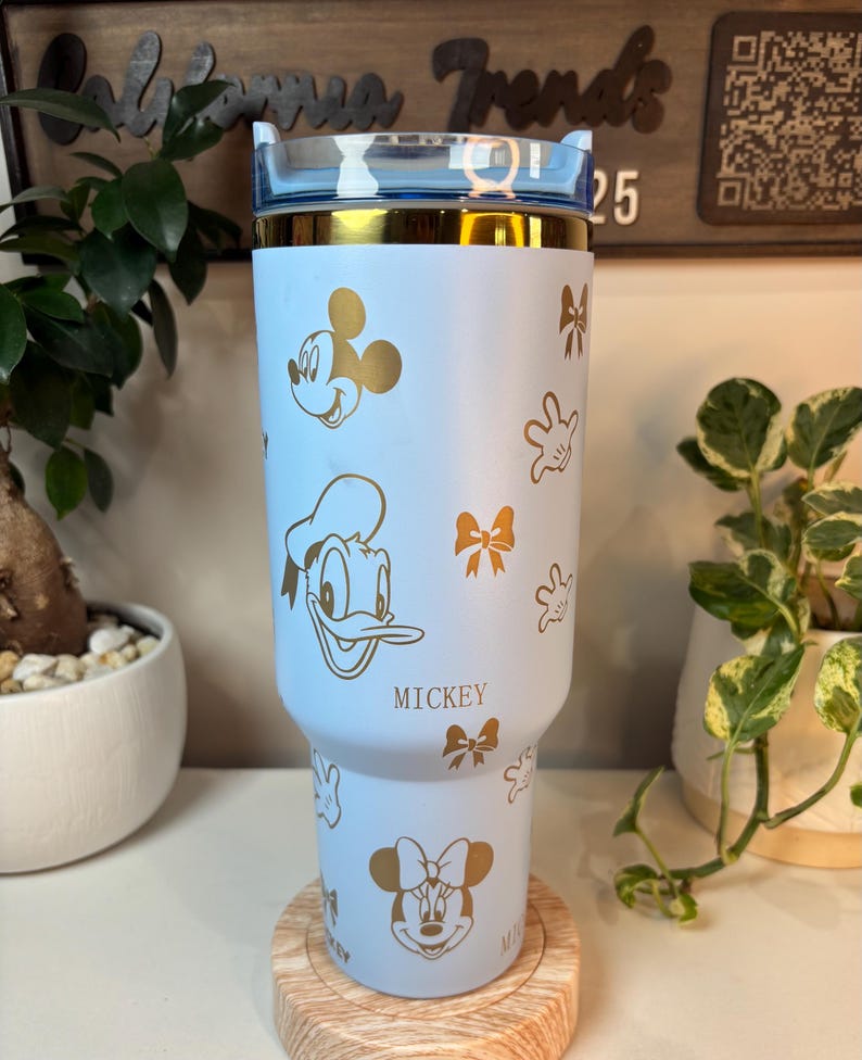 Puede incluir: Un vaso azul claro con dise&ntilde;os dorados de Mickey Mouse, Donald Duck y Minnie Mouse, junto con lazos y guantes. El vaso tiene un borde dorado y una tapa azul transparente. La palabra "Mickey" est&aacute; impresa.