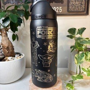 May include: Black Owala water bottle featuring a silver "May the Force Be With You" design and Star Wars character illustrations. The bottle sits on a light brown coaster, with plants in the background.