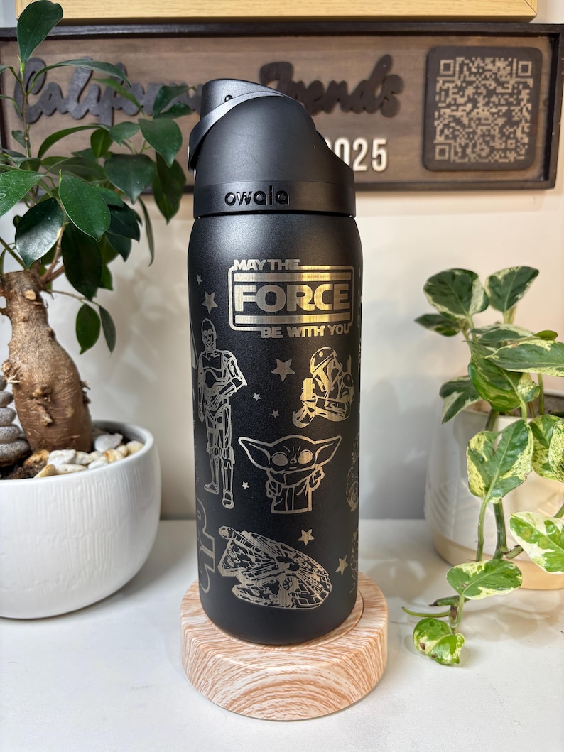 May include: Black Owala water bottle featuring a silver "May the Force Be With You" design and Star Wars character illustrations. The bottle sits on a light brown coaster, with plants in the background.