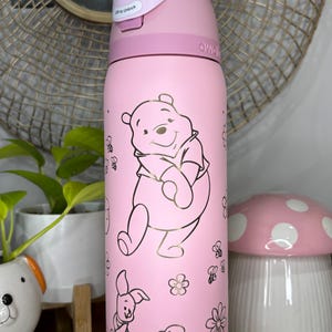 May include: Pink water bottle featuring Winnie the Pooh and Piglet illustrations with bees and flowers. The bottle has a light pink lid and a push-button opening. The text "Lift to Unlock" is visible on the lid.