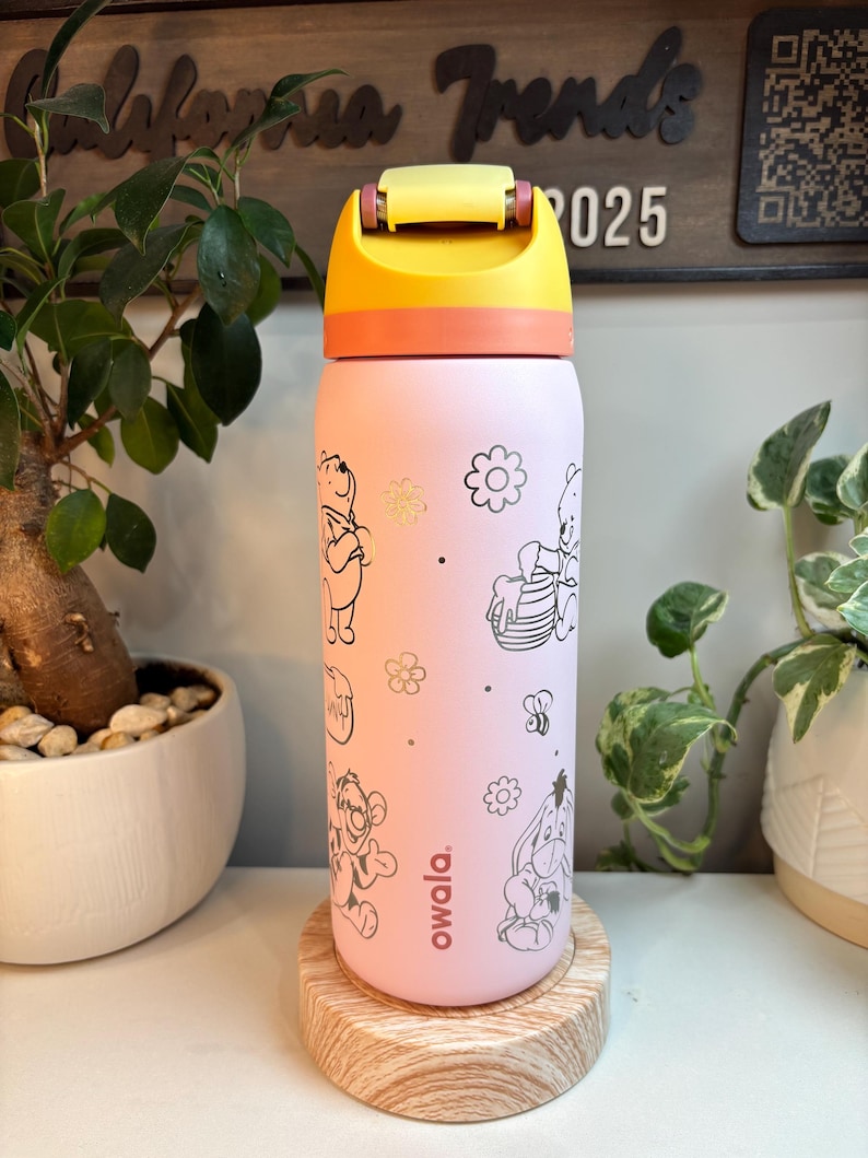Op de afbeelding: Een lichtroze Owala-waterfles met zwarte lijntekeningen van Winnie de Poeh-personages, waaronder Teigetje, Iejoor en Poeh met een honingpot, samen met bloemen en bijen. De fles heeft een gele en oranje dop.