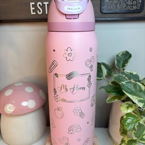 May include: A light pink water bottle with a flip-top lid featuring the text "Lift to Unlock". The bottle is decorated with gold-colored line art icons including pencils, scissors, apples, rainbows, and a central rectangular label reading "Ms Flores".