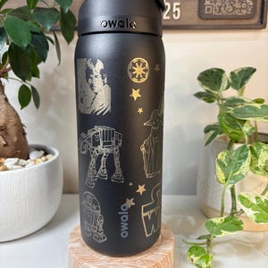 May include: Black Owala water bottle with engraved Star Wars characters and symbols. The bottle features Luke Skywalker, an AT-AT, R2-D2, Yoda, and the Star Wars logo. The bottle is on a wooden coaster.