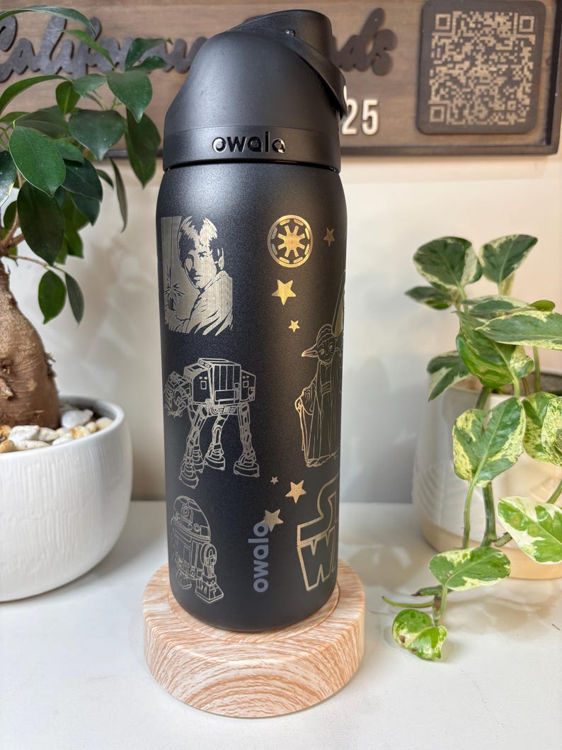May include: Black Owala water bottle with engraved Star Wars characters and symbols. The bottle features Luke Skywalker, an AT-AT, R2-D2, Yoda, and the Star Wars logo. The bottle is on a wooden coaster.
