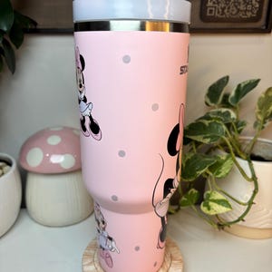 Puede incluir: Un vaso de acero inoxidable rosa con ilustraciones de Minnie Mouse y lunares grises. El vaso tiene una tapa blanca y una parte superior rosa. Est&aacute; sobre un posavasos de madera.
