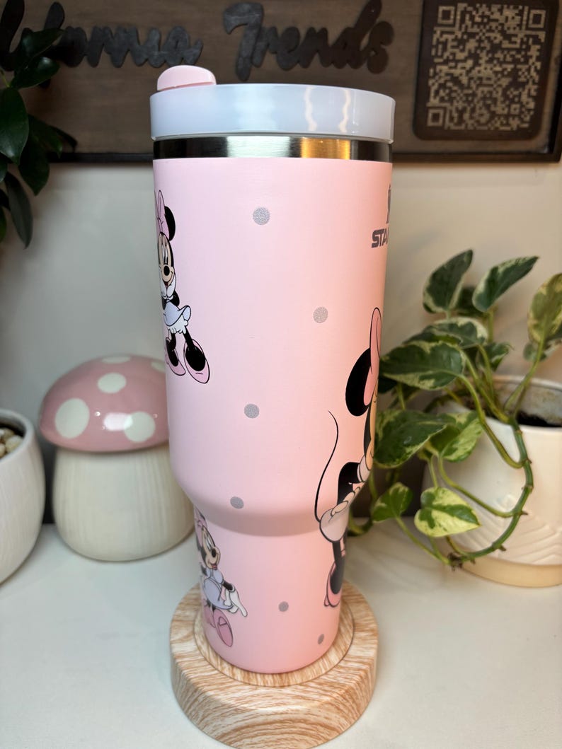 Puede incluir: Un vaso de acero inoxidable rosa con ilustraciones de Minnie Mouse y lunares grises. El vaso tiene una tapa blanca y una parte superior rosa. Est&aacute; sobre un posavasos de madera.