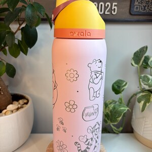 Op de afbeelding: Een lichtroze Owala-waterfles met een geel en oranje deksel en een draaglus. De fles is versierd met zwarte lijntekeningen van Winnie de Poeh, Teigetje, een honingpot met het label Hunnny, kleine bloemen en bijtjes.
