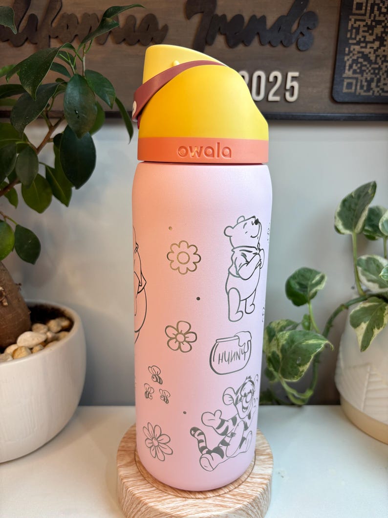 Op de afbeelding: Een lichtroze Owala-waterfles met een geel en oranje deksel en een draaglus. De fles is versierd met zwarte lijntekeningen van Winnie de Poeh, Teigetje, een honingpot met het label Hunnny, kleine bloemen en bijtjes.