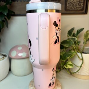 Puede incluir: Un vaso t&eacute;rmico rosa con ilustraciones de Minnie Mouse y lunares. El vaso tiene un asa rosa claro y una tapa blanca y rosa claro. Est&aacute; sobre un posavasos de madera.
