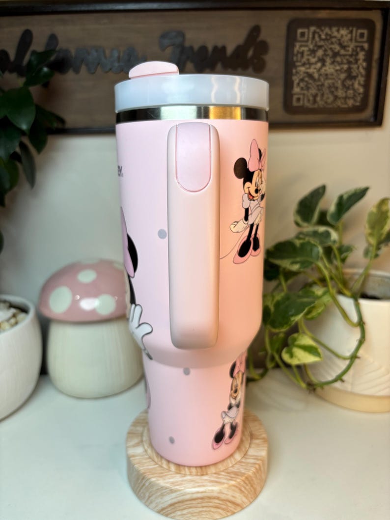 Puede incluir: Un vaso t&eacute;rmico rosa con ilustraciones de Minnie Mouse y lunares. El vaso tiene un asa rosa claro y una tapa blanca y rosa claro. Est&aacute; sobre un posavasos de madera.