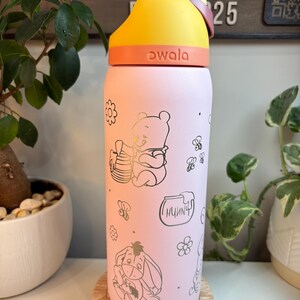 Op de afbeelding: Een lichtroze Owala-waterfles met een gele en oranje dop. De fles is versierd met goudkleurige lijntekeningen van Winnie de Poeh die honing eet, Iejoor, bijen, bloemen en een honingpot met het woord Hunny erop geschreven.