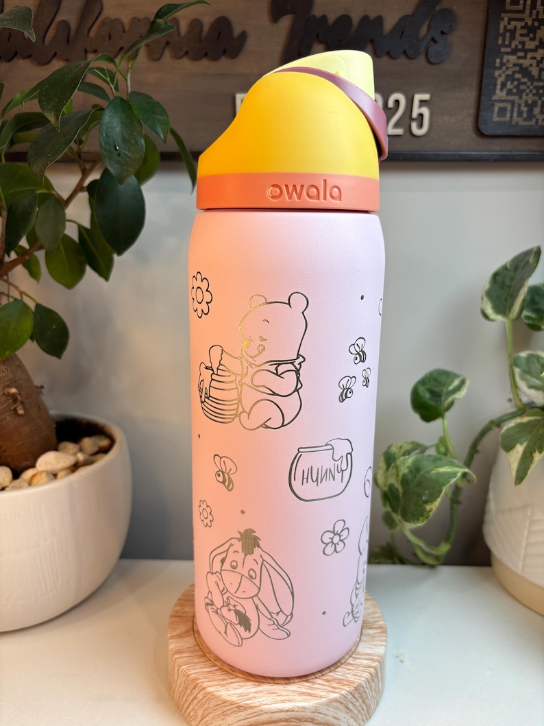 Op de afbeelding: Een lichtroze Owala-waterfles met een gele en oranje dop. De fles is versierd met goudkleurige lijntekeningen van Winnie de Poeh die honing eet, Iejoor, bijen, bloemen en een honingpot met het woord Hunny erop geschreven.