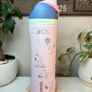 May include: A pink Owala water bottle featuring Snoopy illustrations. The bottle has a blue, green, and pink lid. The bottle is sitting on a wooden coaster. The background includes plants and a wooden sign.