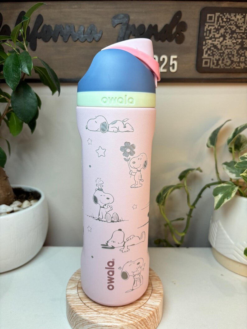 May include: A pink Owala water bottle featuring Snoopy illustrations. The bottle has a blue, green, and pink lid. The bottle is sitting on a wooden coaster. The background includes plants and a wooden sign.