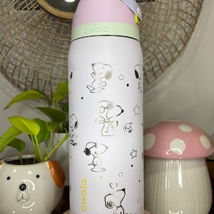Puede incluir: Una botella de agua blanca Owala con ilustraciones de Snoopy y estrellas. La botella tiene una tapa rosa y verde con un asa gris. El logotipo de Owala est&aacute; impreso en amarillo. Un macetero de cer&aacute;mica con forma de perro y una decoraci&oacute;n con forma de hongo est&aacute;n en el fondo.
