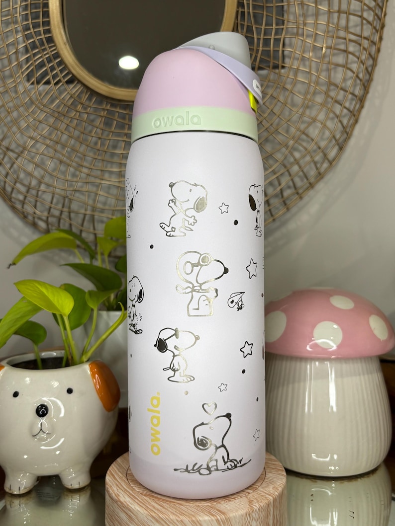 Puede incluir: Una botella de agua blanca Owala con ilustraciones de Snoopy y estrellas. La botella tiene una tapa rosa y verde con un asa gris. El logotipo de Owala est&aacute; impreso en amarillo. Un macetero de cer&aacute;mica con forma de perro y una decoraci&oacute;n con forma de hongo est&aacute;n en el fondo.