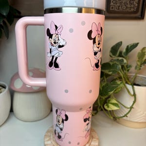 Puede incluir: Un vaso t&eacute;rmico rosa con ilustraciones de Minnie Mouse y lunares grises. El vaso tiene un asa, una tapa blanca y plateada y un asa rosa. El vaso est&aacute; sobre un posavasos de madera.