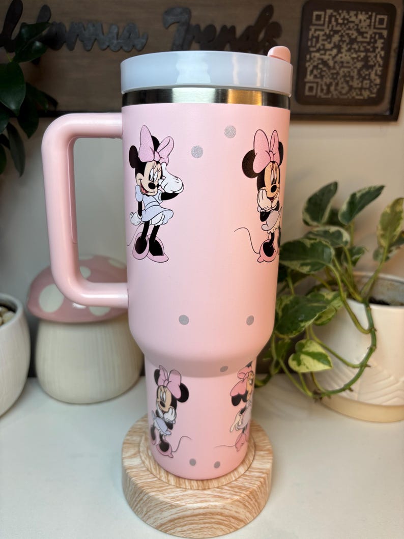 Puede incluir: Un vaso t&eacute;rmico rosa con ilustraciones de Minnie Mouse y lunares grises. El vaso tiene un asa, una tapa blanca y plateada y un asa rosa. El vaso est&aacute; sobre un posavasos de madera.