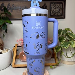 May include: A light purple Stanley tumbler featuring Snoopy illustrations in gold. The tumbler has a handle, a white and silver rim, and a light blue bow on the lid. The tumbler is sitting on a wooden coaster.