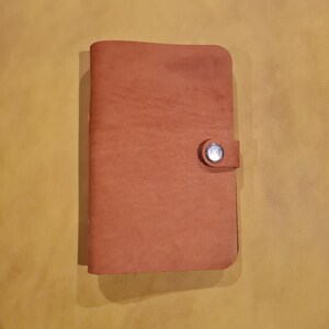 Op de afbeelding: Een handgemaakt, roodbruin leren notitieboek met een knoopsluiting. Het notitieboek is rechthoekig met afgeronde hoeken en een getextureerd oppervlak. De knop is zilverkleurig en rond, wat een veilige sluiting biedt.