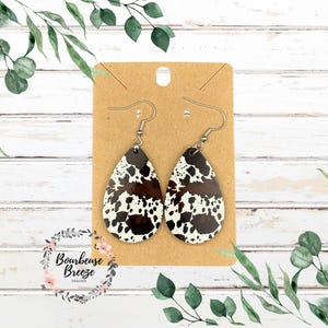 May include: A pair of teardrop-shaped earrings featuring a brown and white cow print pattern. The earrings are attached to a rectangular tan display card with a hole at the top for hanging. Each earring includes a silver-toned metal hook wire.