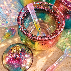 May include: A round resin jar with a matching lid, featuring a multi-coloured gradient of pink, blue, and yellow infused with iridescent glitter. The jar includes a small matching spoon, with additional jars and spoons visible nearby.