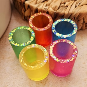 May include: Five brightly coloured resin shot glasses. Each glass is a different colour: green, orange, blue, yellow, and pink. The rims are decorated with glitter. The shot glasses are cylindrical and appear handmade.