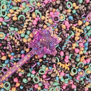 May include: A purple star-shaped wand with glitter rests on a colourful sensory bin. The bin is filled with black beans, pink, teal, and purple rings, and orange and yellow spiral pasta. The sensory bin is designed for children.