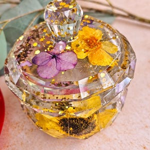 May include: A clear, faceted resin jar with a matching lid featuring a diamond-shaped handle. The jar is embedded with dried yellow flowers, a purple hydrangea petal, and gold glitter flakes throughout the transparent material.