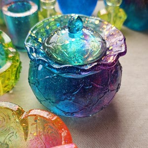 May include: A small, round, lidded container with a shimmering, iridescent finish. The container transitions from blue to purple, with a matching lid and a small, rounded knob. The lid's edges have a ruffled design.