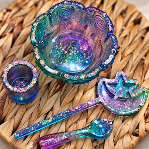 May include: A set of resin items including a scalloped bowl, a small cylindrical cup, a star and crescent moon shaped wand, and a spoon. Each piece features a translucent gradient of blue, purple, and teal with embedded multicoloured glitter.