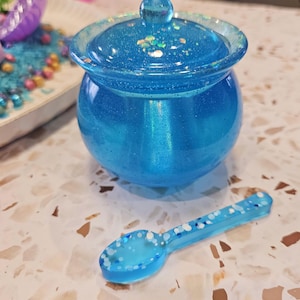 May include: A small blue glass jar with a lid, filled with iridescent glitter. The jar is sitting on a white surface with a matching blue spoon.