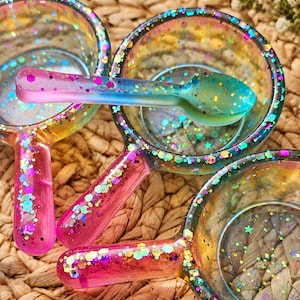 May include: Three small, round, translucent resin bowls with handles, each featuring a vibrant gradient of pink, yellow, and blue colours. The bowls are embedded with multicoloured glitter in various shapes, including stars and hexagons.